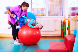 little boy with disability has musculoskeletal therapy by doing exercises in body fixing belts on fit ball